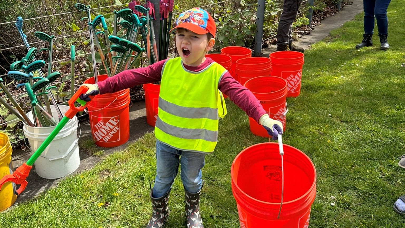 A young boy in a reflective vest holds out an orange bucket and trash grabbing tool.